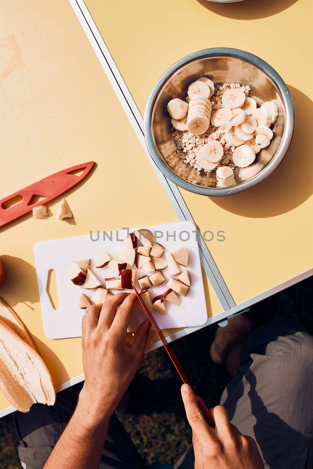 Man preparing healthy breakfast with fruits and oat flakes outdoors on camping. Close up of male hands cutting apples and bananas for breakfast. Camp life