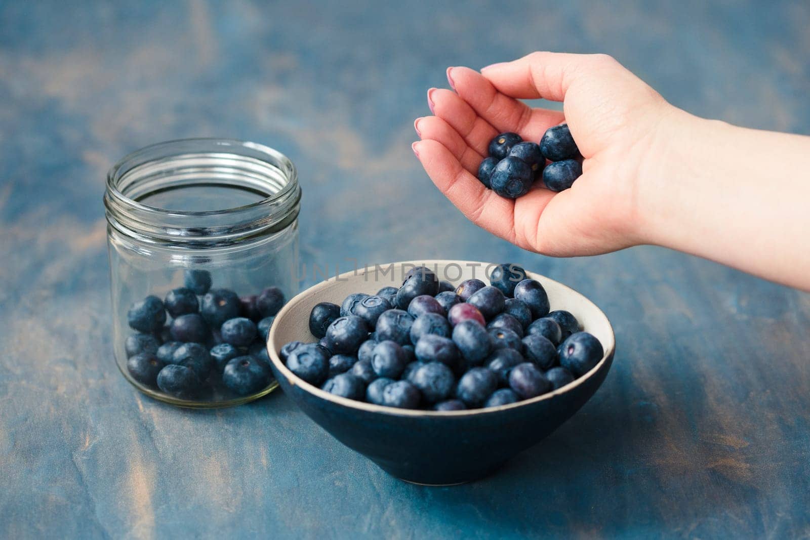 Woman putting freshly gathered blueberries from a jar into a small bowl. Table painted in blue in the background