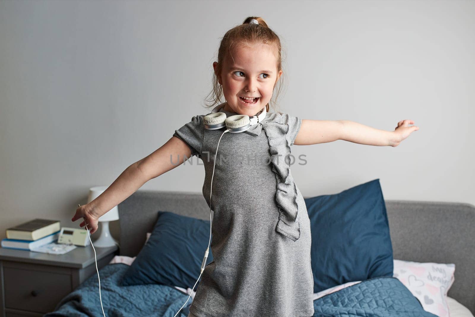 Little girl singing holding headphones cord imitating herself a real singer. Child having fun jumping dancing listening to music on bed in bedroom at home