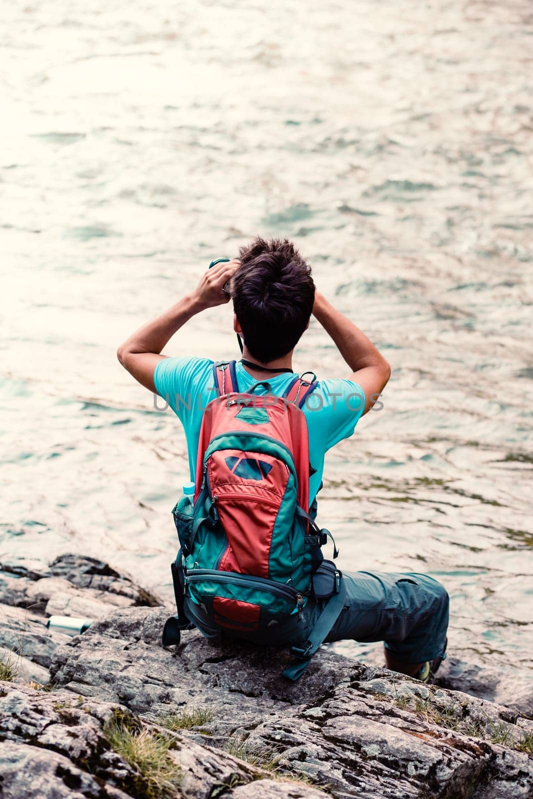 Young tourist with backpack looks through a binoculars, sits on a rock over a river. Boy spends a vacation on wandering with backpack, he is wearing sports summer clothes