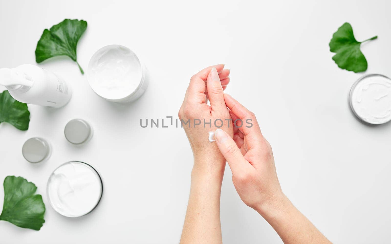 Woman applying  cosmetic moisturizing hand cream. Cosmetic products, green leaves on white table. Spa, manicure, skin care concept. Flat lay, overhead view