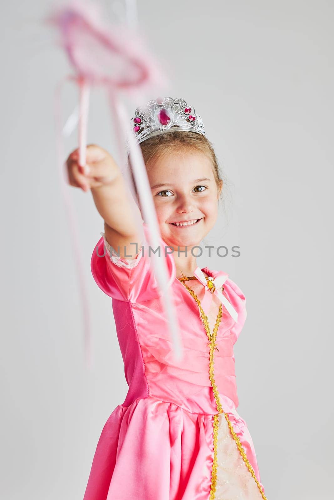 Little girl enjoying her role of princess. Adorable cute 5-6 years old girl wearing pink princess dress and tiara making magic using magic wand standing on plain background