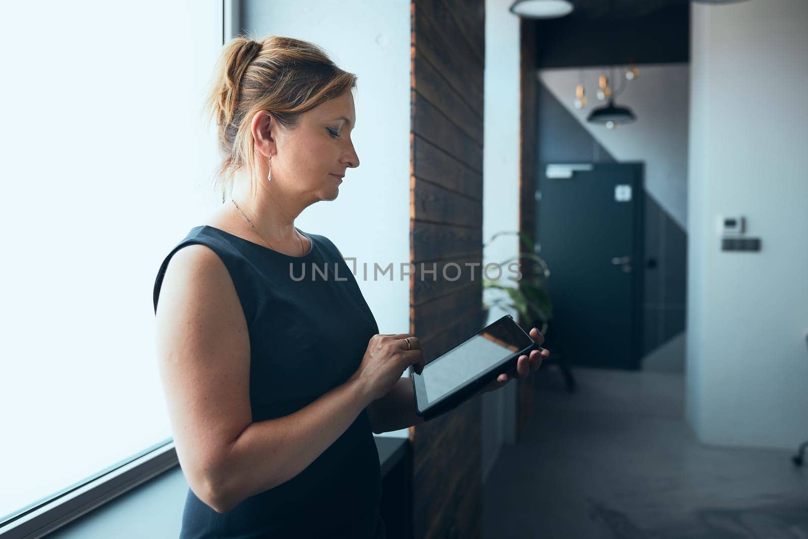 Businesswoman working on tablet in office. Mature woman using touch pad computer standing by window in modern interior. Female manager focused on work holding digital device. Using technology. Open office. Copy space right