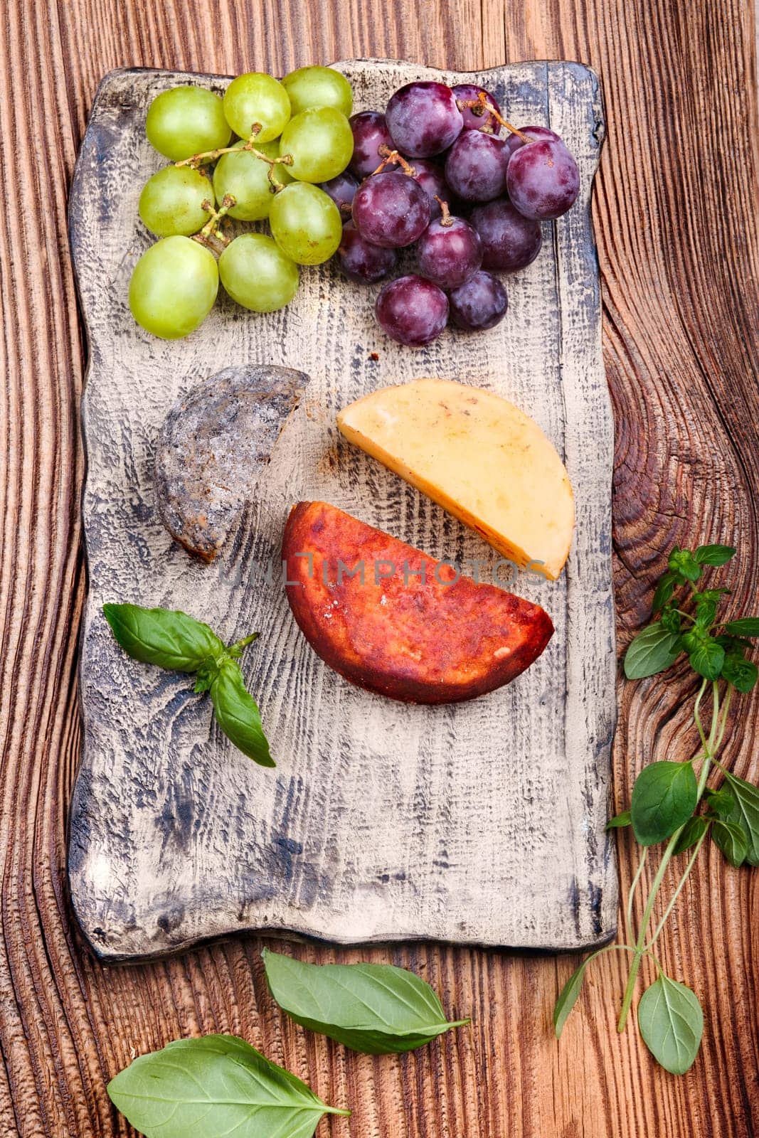Cheese and grapes decorated with basil leaves on handmade pottery plate on old wooden table from above