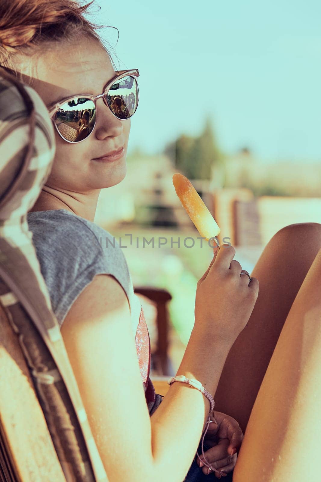 Young woman eating a ice cream, sitting in a chair outdoors on a patio during summer day. Real people, authentic situations