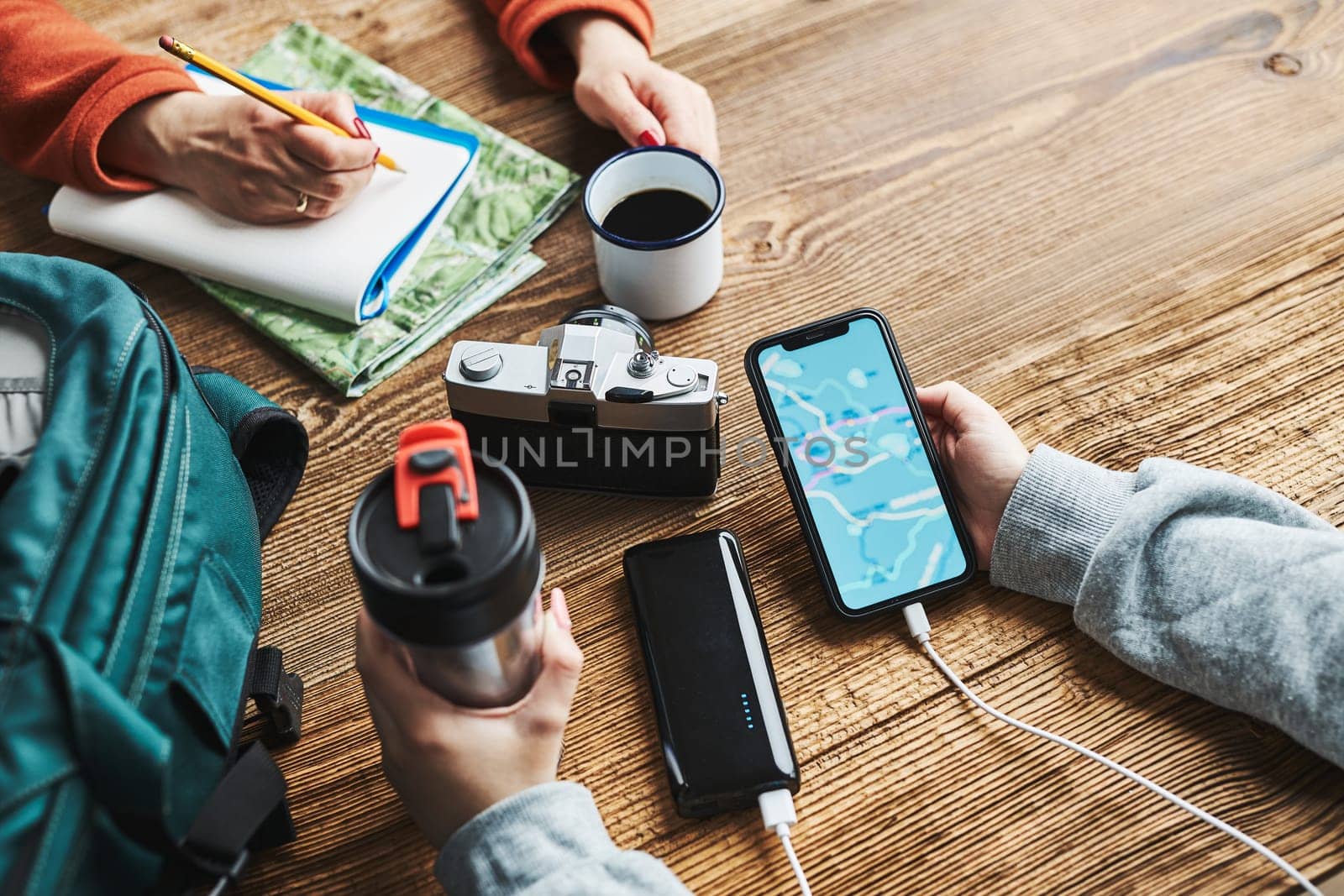 Family planning vacation trip. Girl searching travel destination and routes using navigation map on mobile phone. Woman writing journal and making notes. Charging smartphone with power bank. Using technology while travelling. People sitting at wooden table with camera and backpack during journey. Travel plan concept
