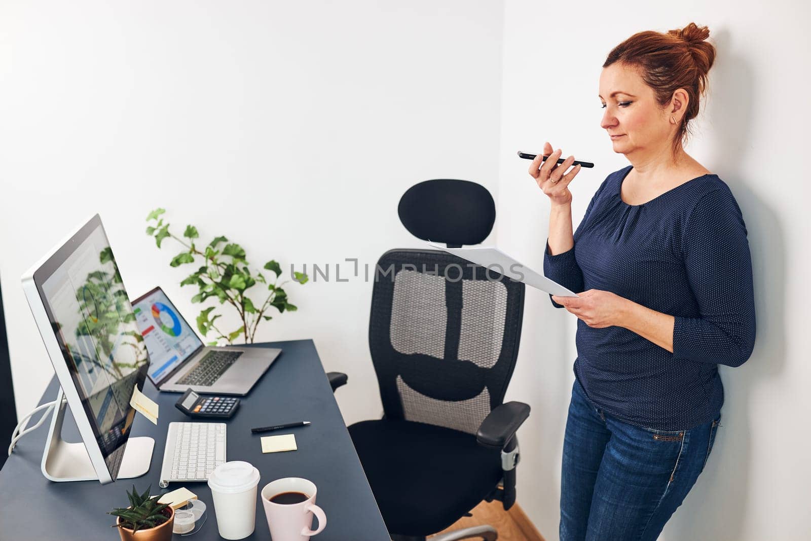 Woman entrepreneur having business  conversation on mobile phone. Businesswoman working in office holding documents and smartphone. Woman standing by desk with computers