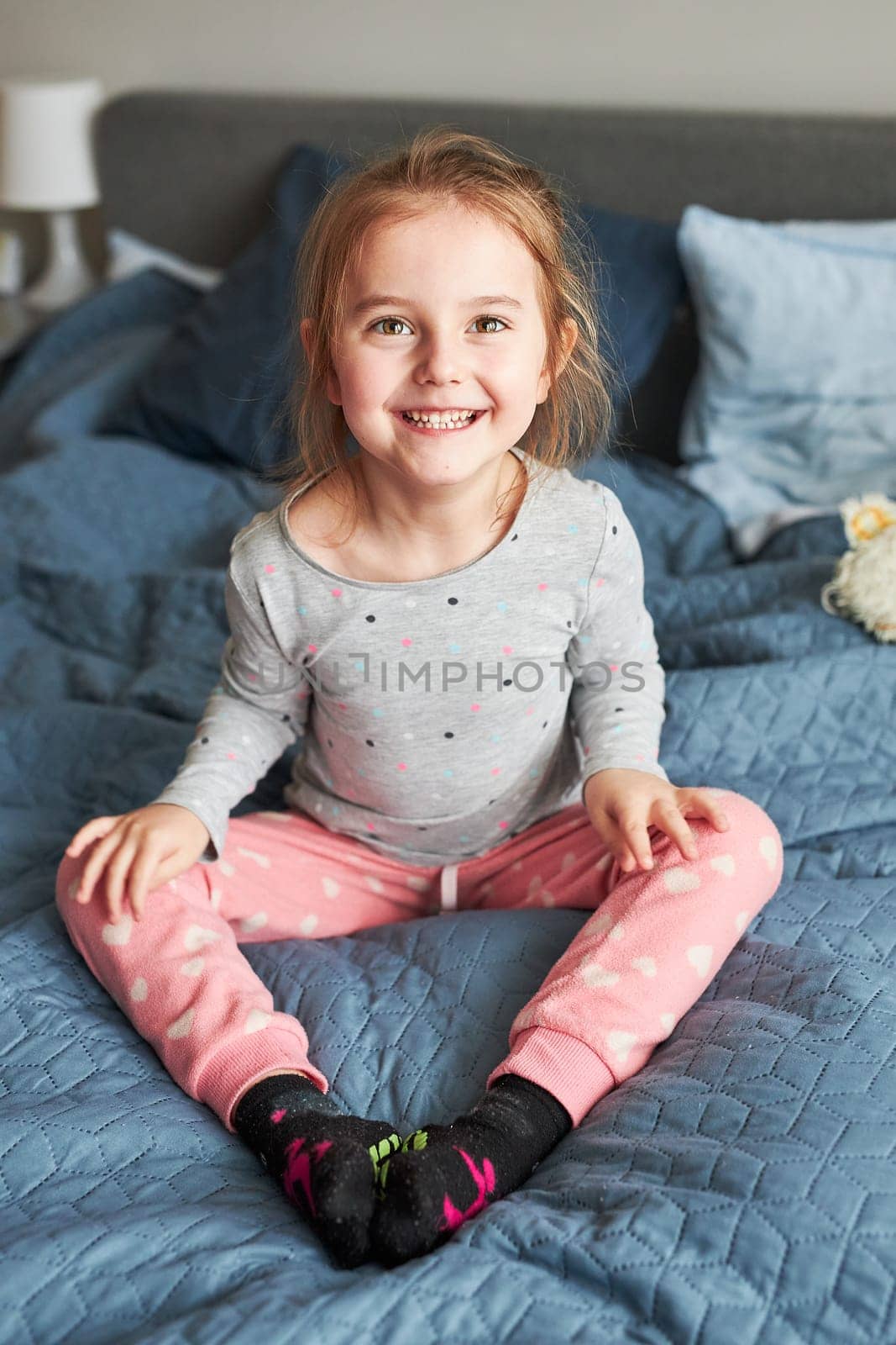 Little happy playful kid girl having fun making silly funny faces jumping on bed in bedroom in the morning at home