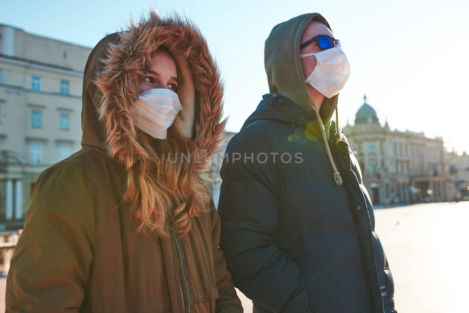 Young woman and man in the hoods wearing the face masks to avoid virus infection and to prevent the spread of disease. People walking in city centre. Virus infection protection. Real people, authentic situations