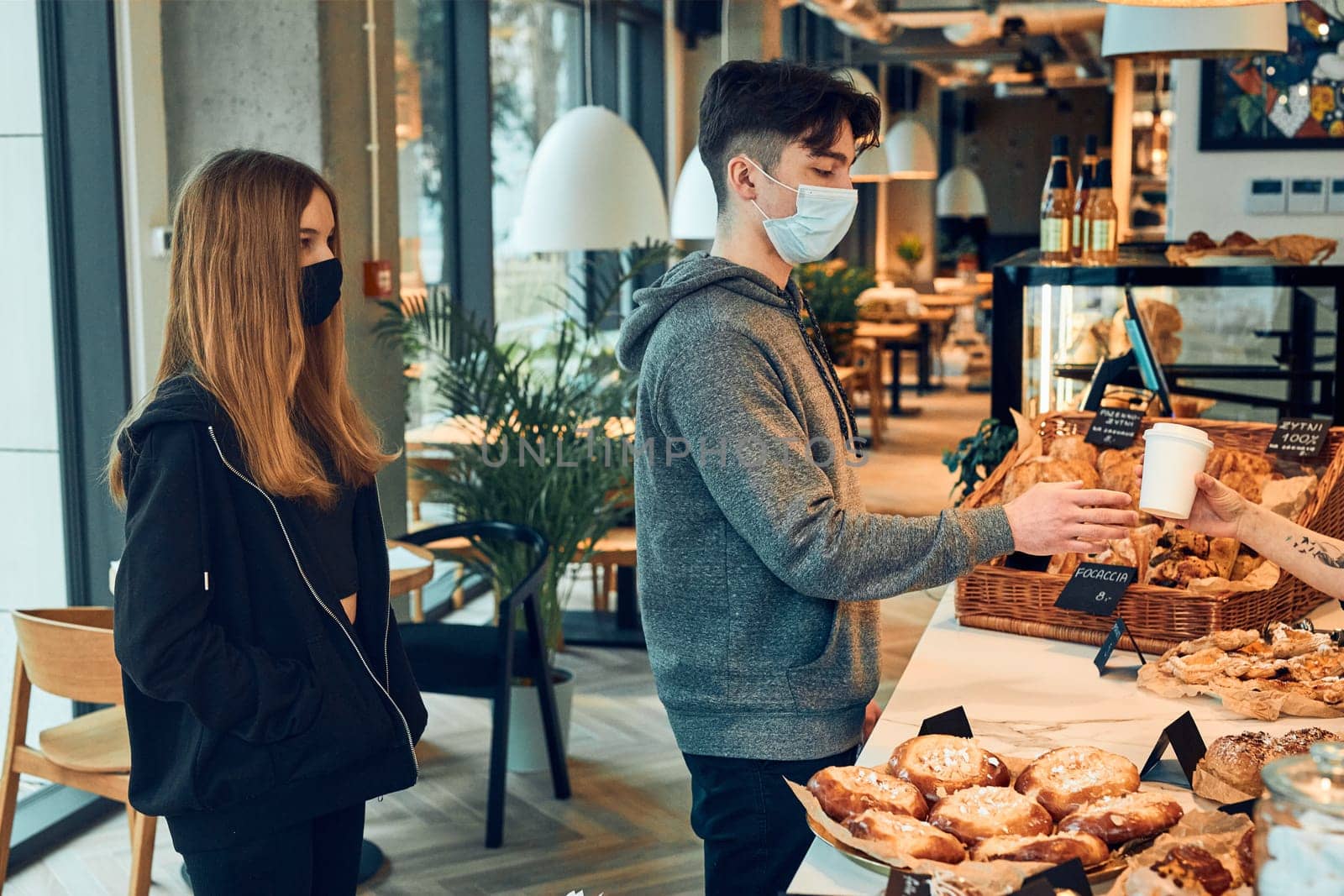People standing in a queue to buy coffee and pastry in the coffee shop to go. Man taking coffee cup from barista. People standing at counter wearing the face masks to avoid virus infection and to prevent the spread of disease in time of coronavirus