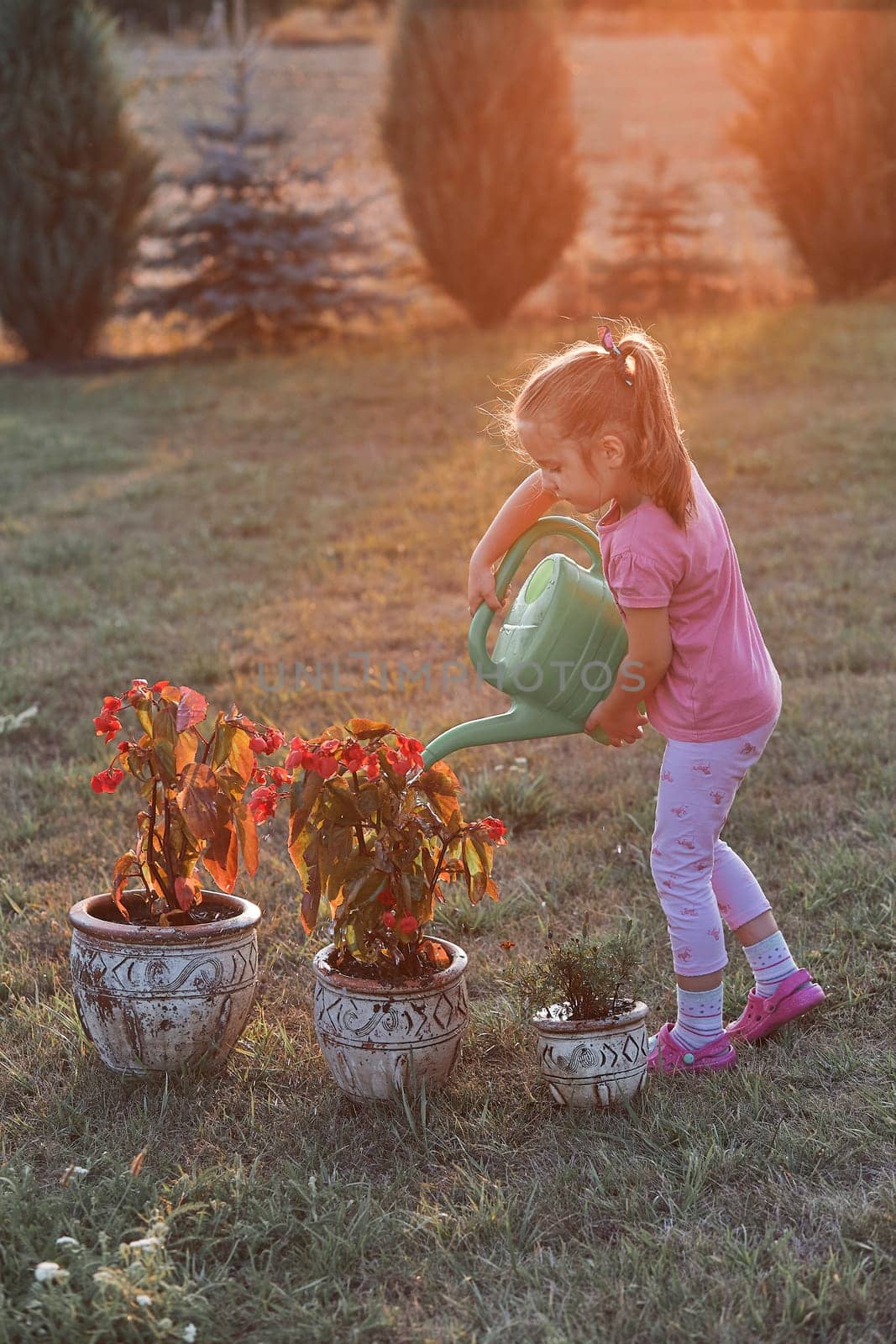 Little girl helping to water the flowers growing in flower pot, pouring water from green watering can, working in backyard at sunset. Candid people, real moments, authentic situations
