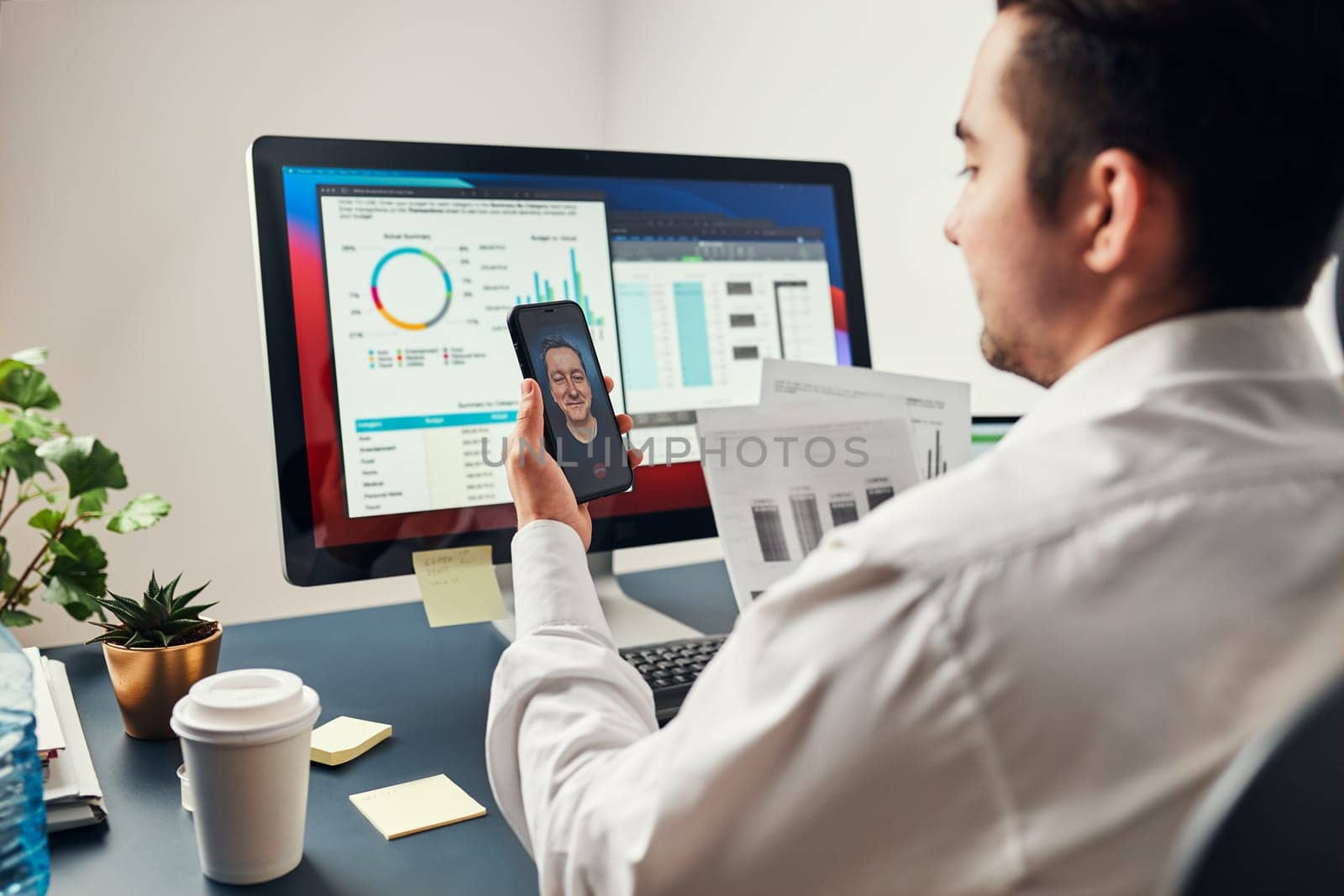 Man entrepreneur having business conversation, video chat on mobile phone. Businessman working with data, charts and graphs on computer screen. Man holding smartphone sitting at desk in office