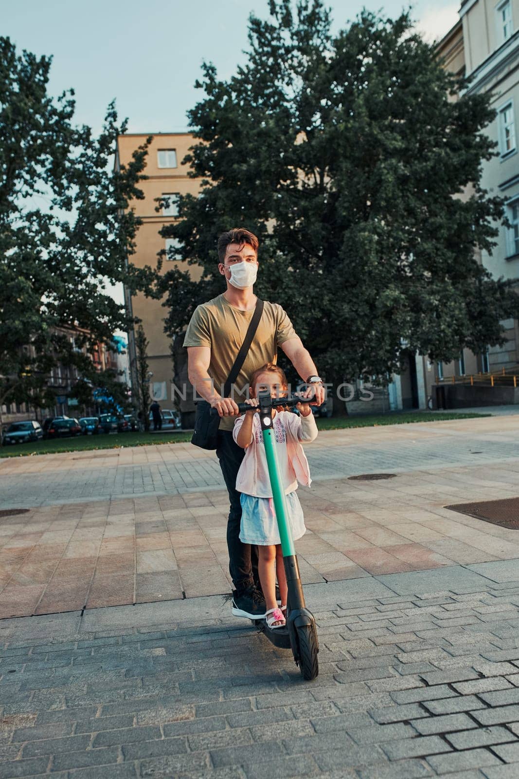 Young man and her sister preschooler riding an electric scooter in the city center. Man wearing the face mask to avoid virus infection and to prevent the spread of disease in time of coronavirus. Candid people, real moments, authentic situation