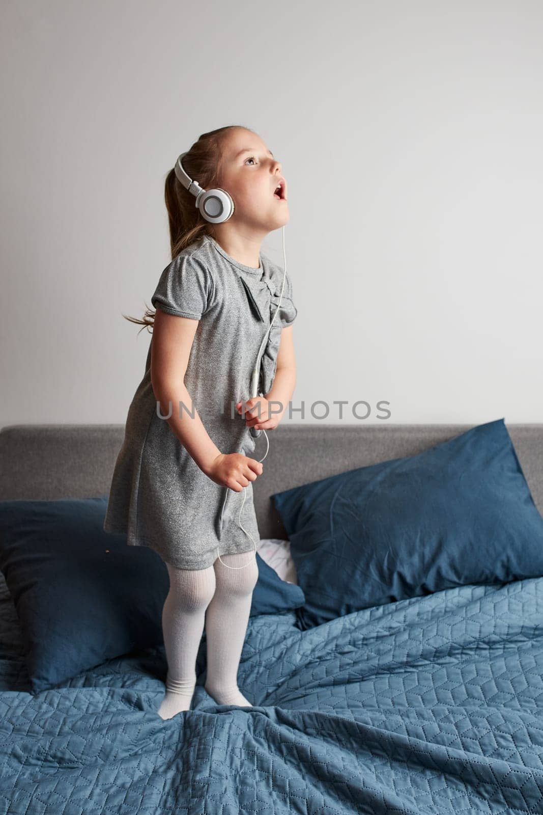 Little girl singing holding headphones cord imitating herself a real singer. Child having fun jumping dancing listening to music on bed in bedroom at home
