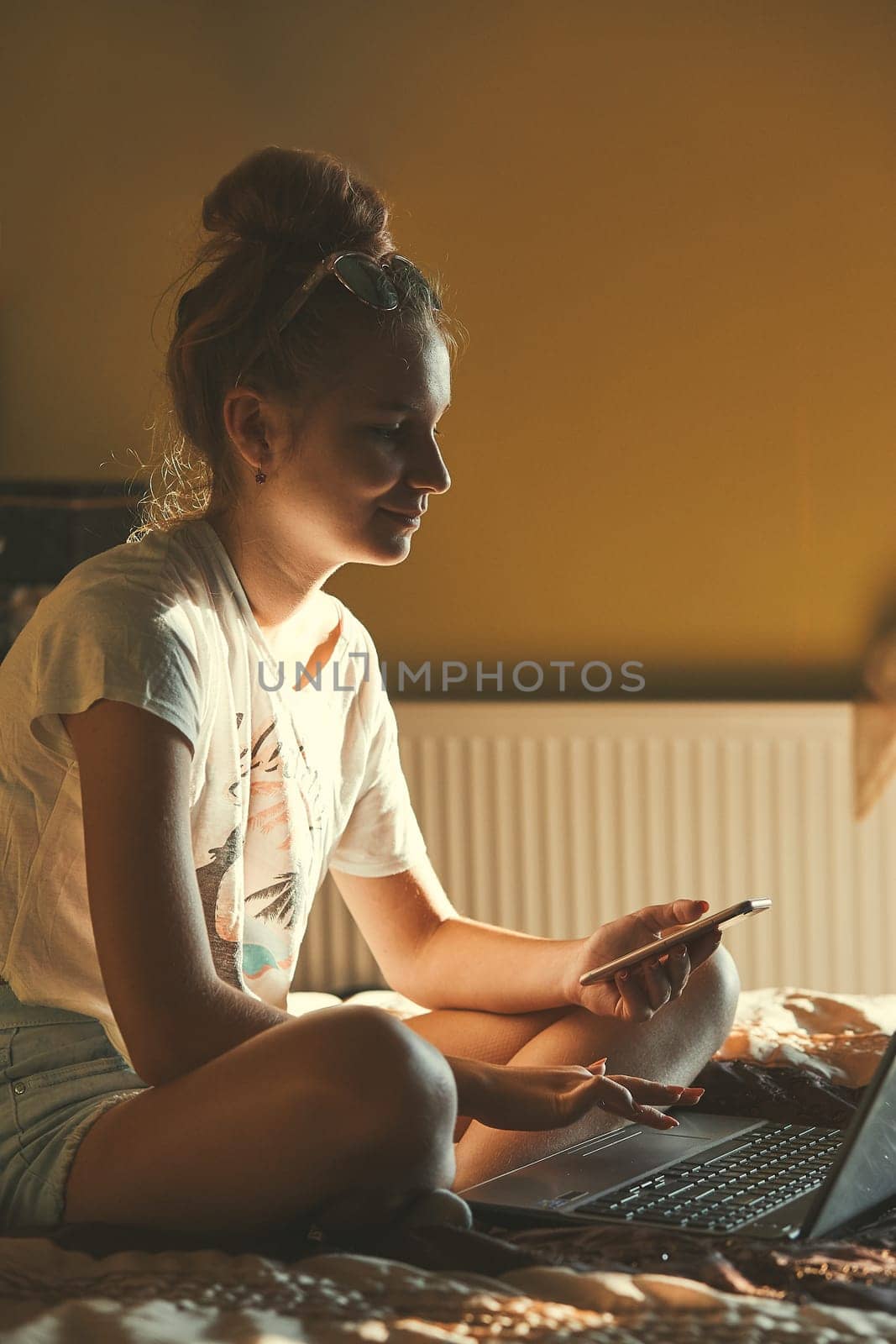 Young woman using portable computer and mobile phone, sitting on a bed, learning online at home. Candid people, real moments, authentic situations