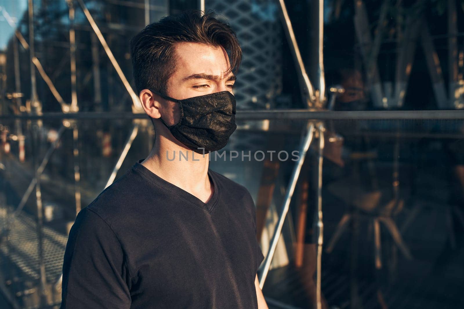Young man walking in the city center along glass store front in the evening, looking away, wearing the face mask to avoid virus infection and to prevent the spread of disease in time of coronavirus