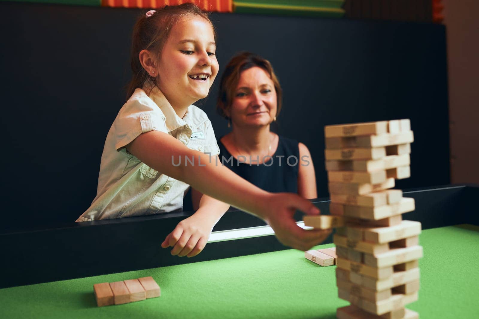 Excited girl playing jenga game with her mom in play room. Girl removing one block from stack and placing it on top of tower. Stacked wooden blocks. Game of skill and fun. Family time