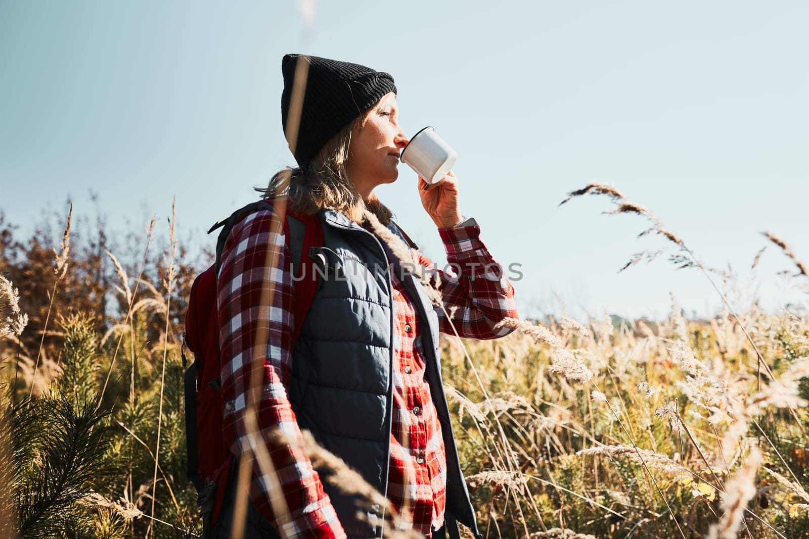 Woman taking break and enjoying the coffee during vacation trip. Woman standing on trail and looking away holding cup of coffee. Woman with backpack hiking through tall grass along path in mountains. Spending summer vacation close to nature