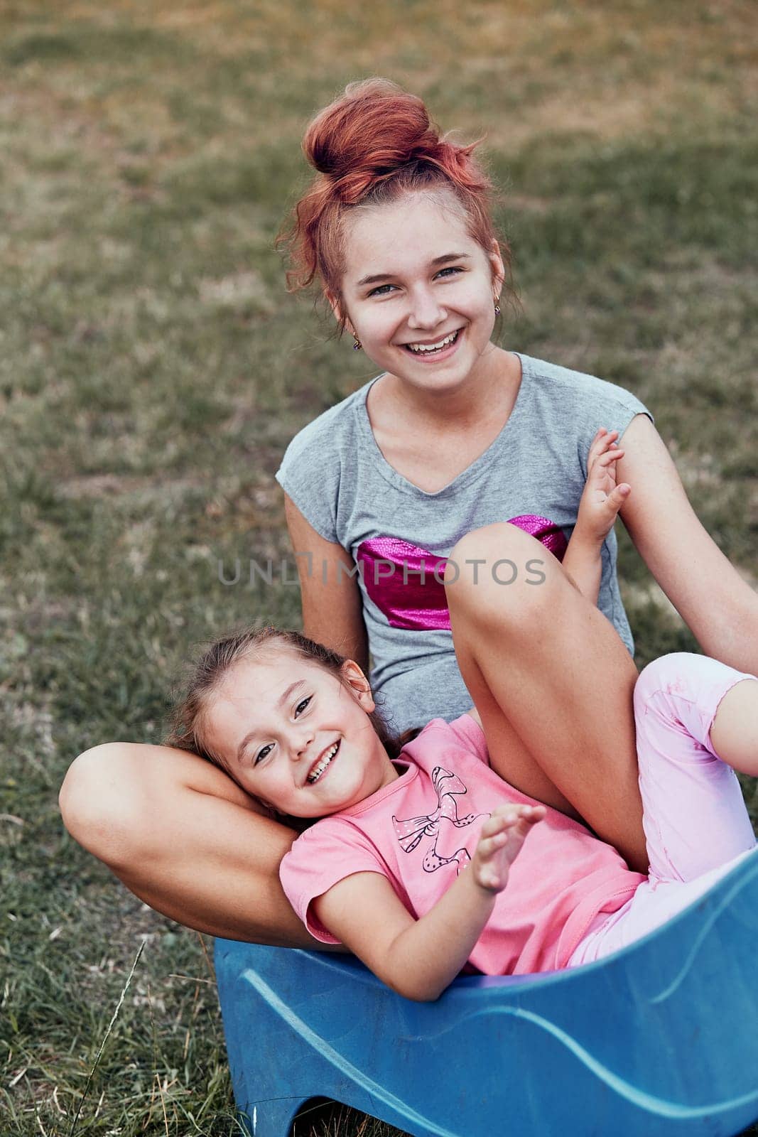 Teenage girl playing with her younger sister in a home playground in a backyard. Happy smiling sisters having fun on a slide together on summer day. Real people, authentic situations