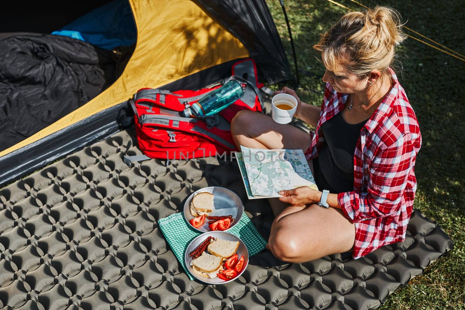 Woman having breakfast and planning day sitting by tent at camping. Female actively spending summer vacations close to nature outdoors. Concept of camp life