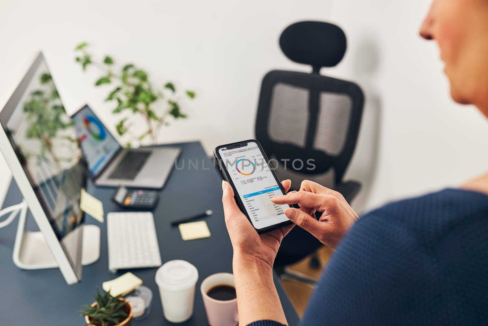 Businesswoman working with financial data on charts and tables on smartphone and computer. Woman entrepreneur looking at sale stats standing by desk in office