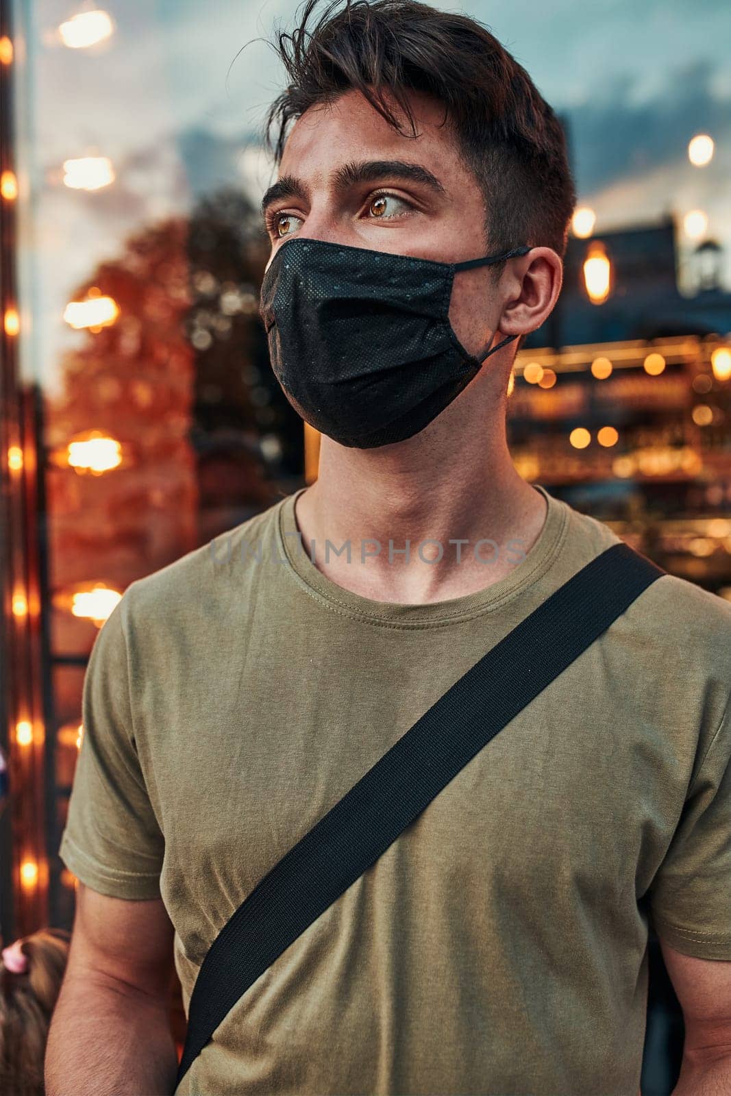 Young man standing at store front in the city center in the evening, looking away, wearing the face mask to avoid virus infection and to prevent the spread of disease in time of coronavirus