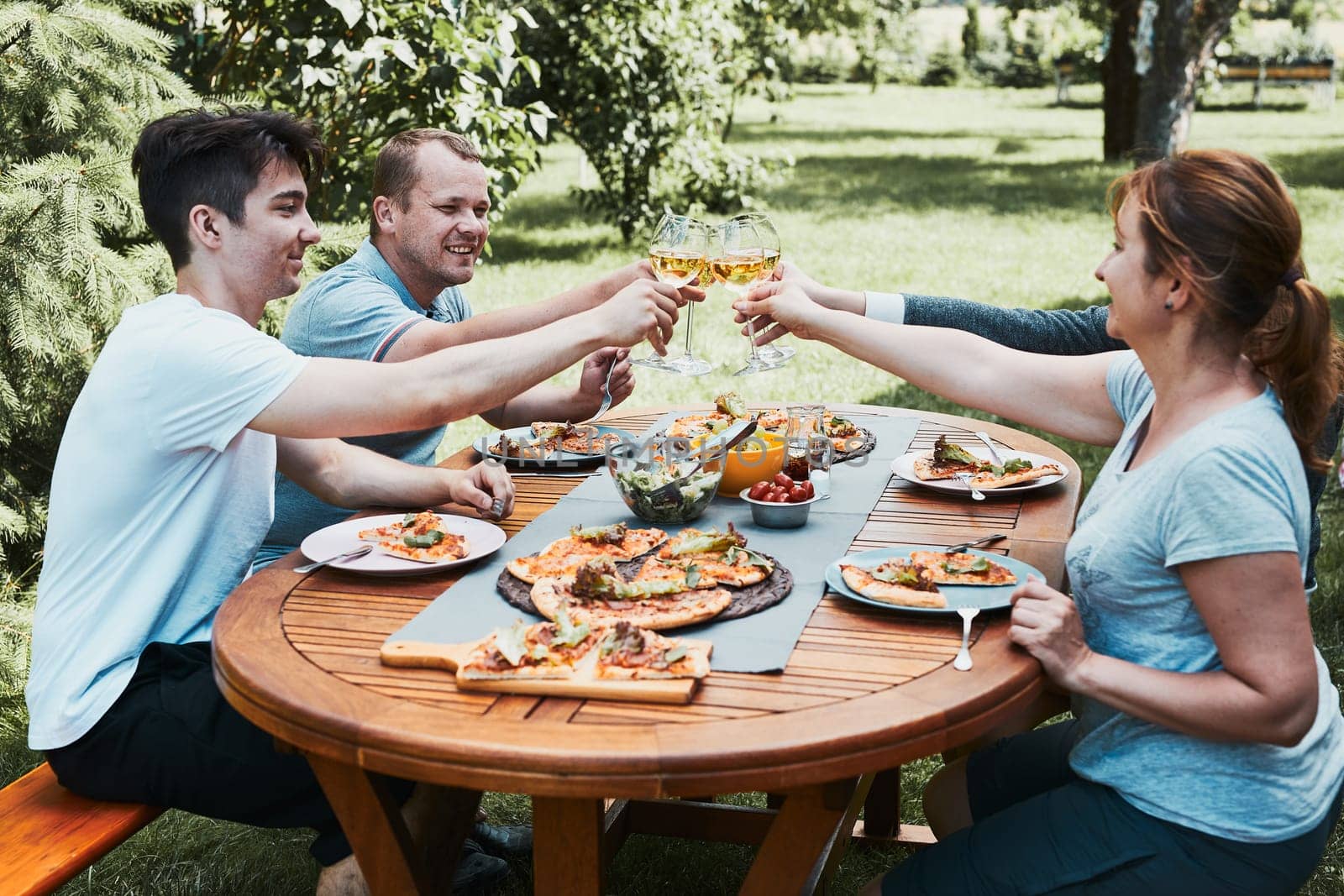 Friends making toast during summer picnic outdoor dinner in a home garden. Close up of people holding wine glasses with white wine over table with pizza, salads and fruits. Dinner in a orchard in a backyard