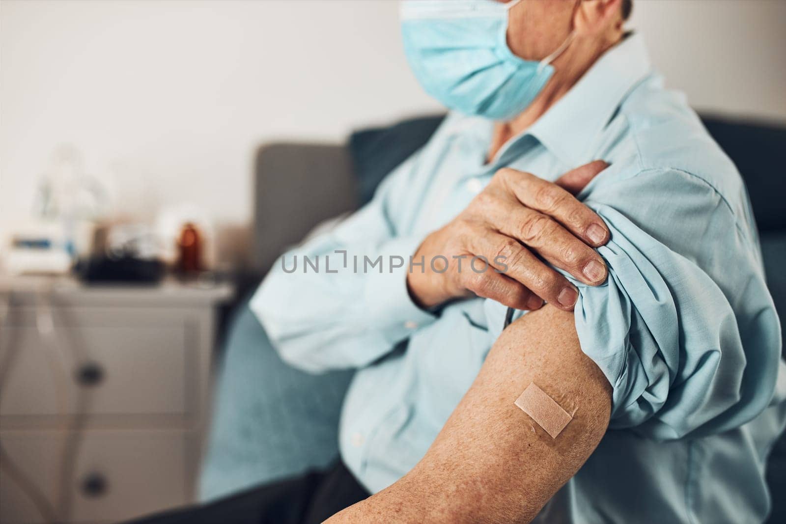 Senior man patient holding shirt sleeve up with a plaster in place of injection of vaccine. Covid-19 or coronavirus vaccination. Man wearing face mask
