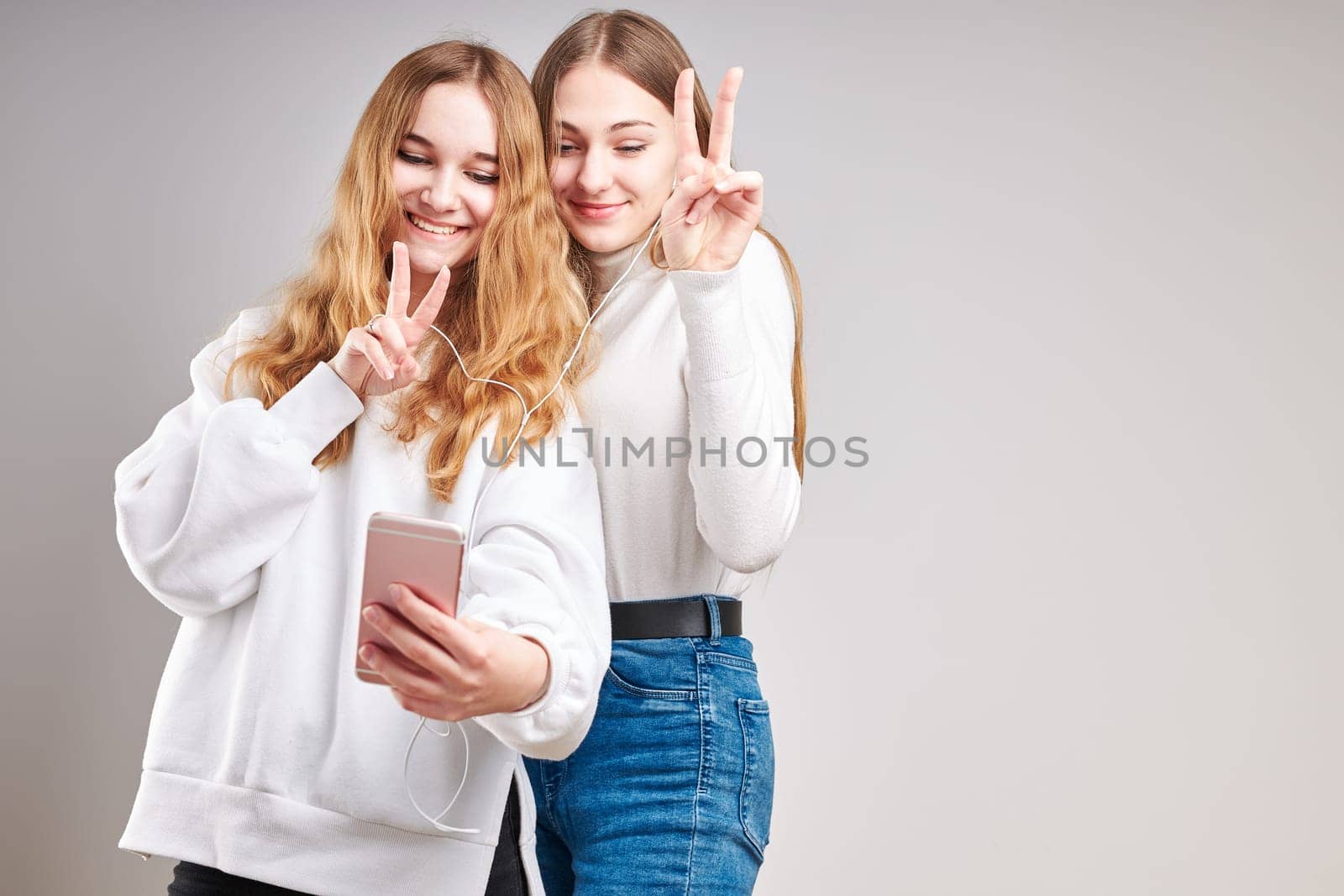 Young women girls listening to music together streaming content having fun watching video enjoying video chat talking with friends making gestures faces using smartphone earphones headphones standing over plain grey background