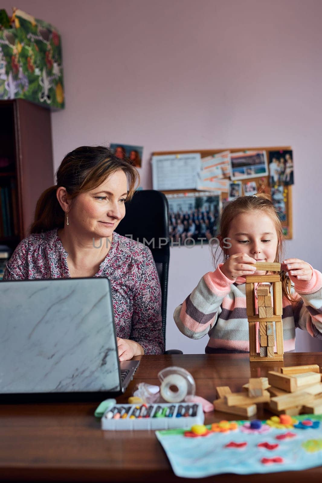 Woman mother working doing her job remotely during video chat call stream online course webinar on laptop from home while her daughter playing with bricks toy. Woman sitting at desk in front of computer looking at screen