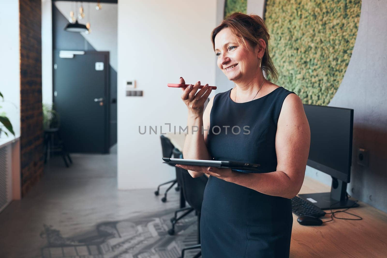 Businesswoman having business call working on tablet in office. Mature busy woman using touch pad computer standing by window in modern interior. Female manager focused on work holding digital device. Using technology