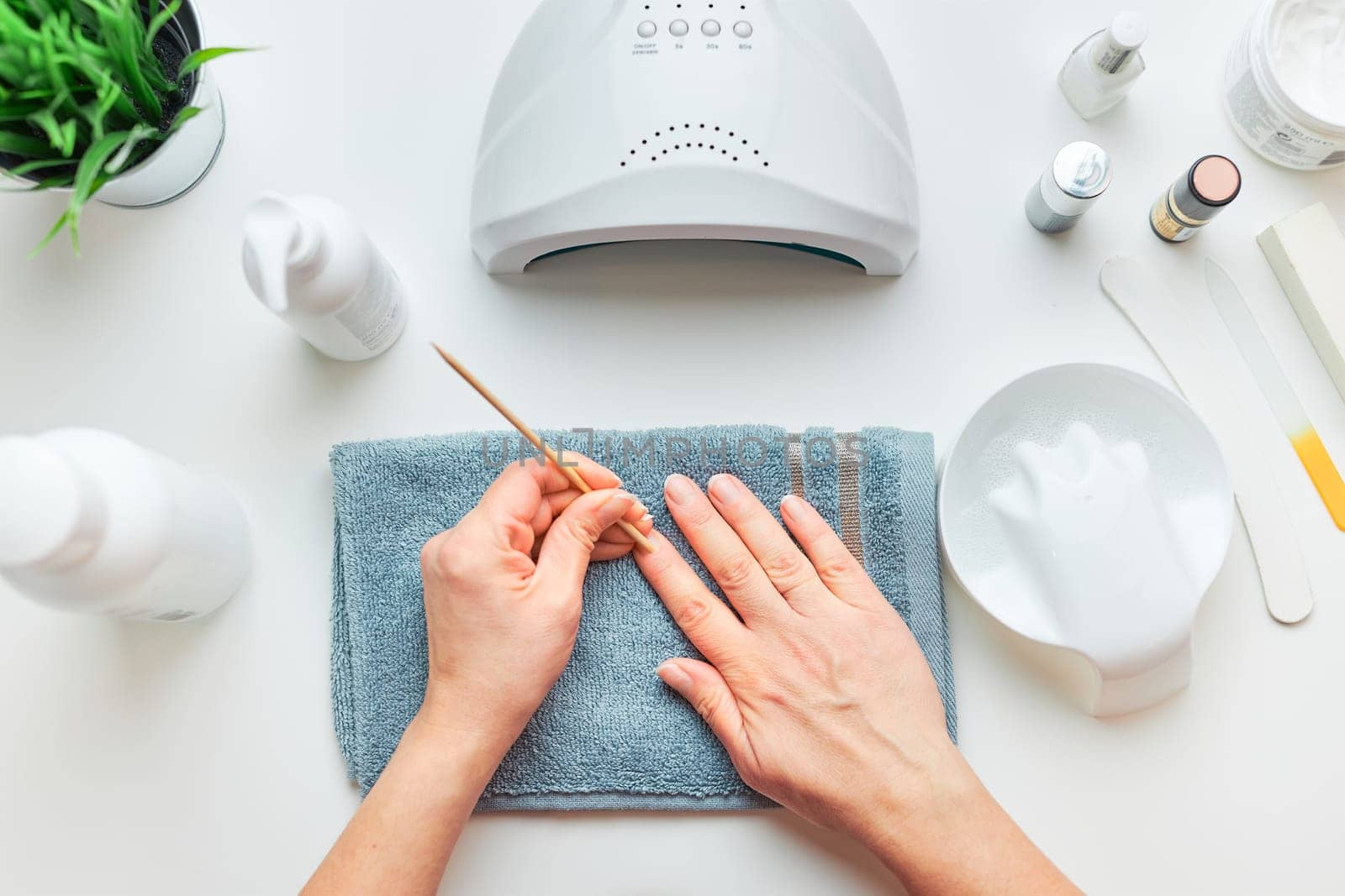Woman preparing nails to apply gel hybrid polish using UV lamp. Beauty wellness spa treatment concept. Cosmetic products, UV lamp, green leaves on white table. Spa, manicure, skin care concept. Flat lay, overhead view