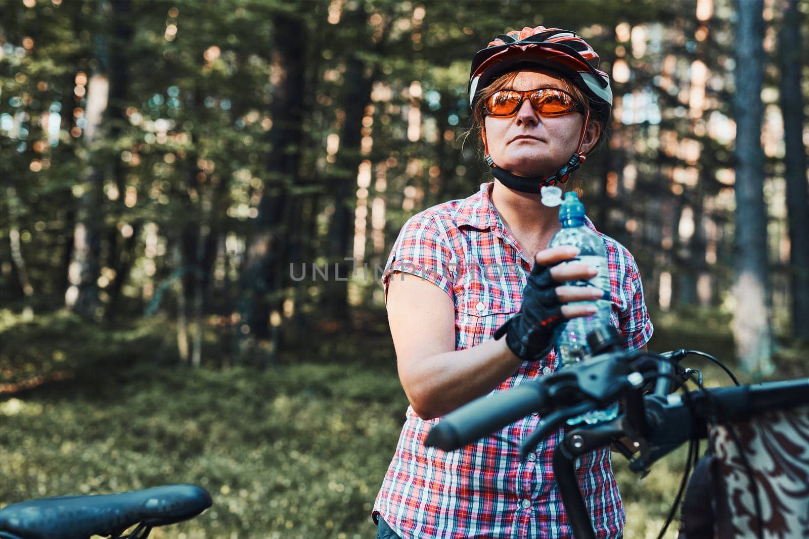 Active woman spending free summer vacation time on a bicycle trip in a forest. Woman wearing bicycle helmet and gloves holding bike with basket and drinking a water from bottle