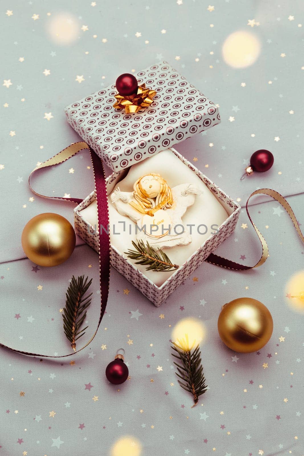 Wrapping Christmas gift into box. Decorating using red ribbon, red Christmas balls and pine twigs. Pink fabric in the background. Blurred Christmas lights. Flat top-down composition. Figure of angel made of earthen with love inscription as Christmas decoration