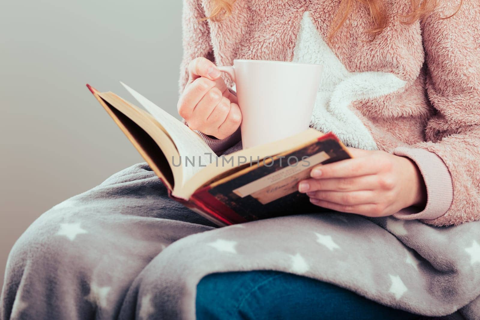 Girl enjoying the reading a book and drinking coffee at home. Young woman sitting on a chair, wrapped in blanket, holding book, relaxing at home. Portrait orientation. Front view