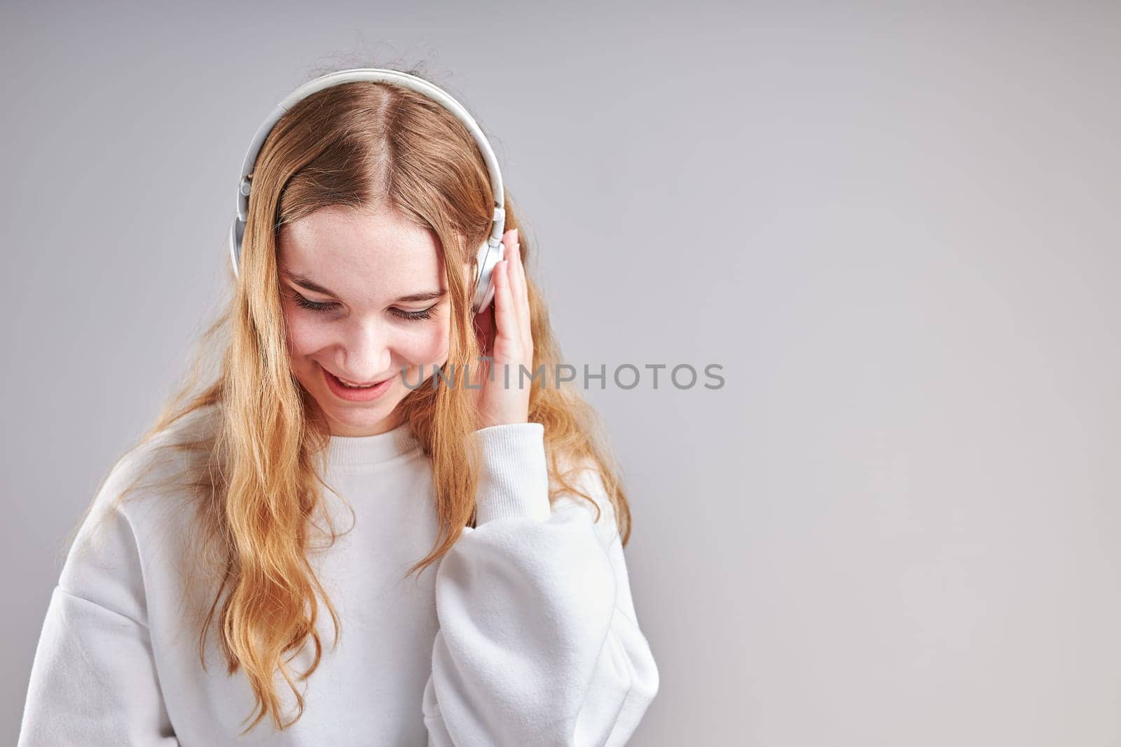 Young woman girl listening to music streaming content having fun watching video enjoying video chat talking with friends making gestures faces using smartphone earphones headphones standing over plain grey background