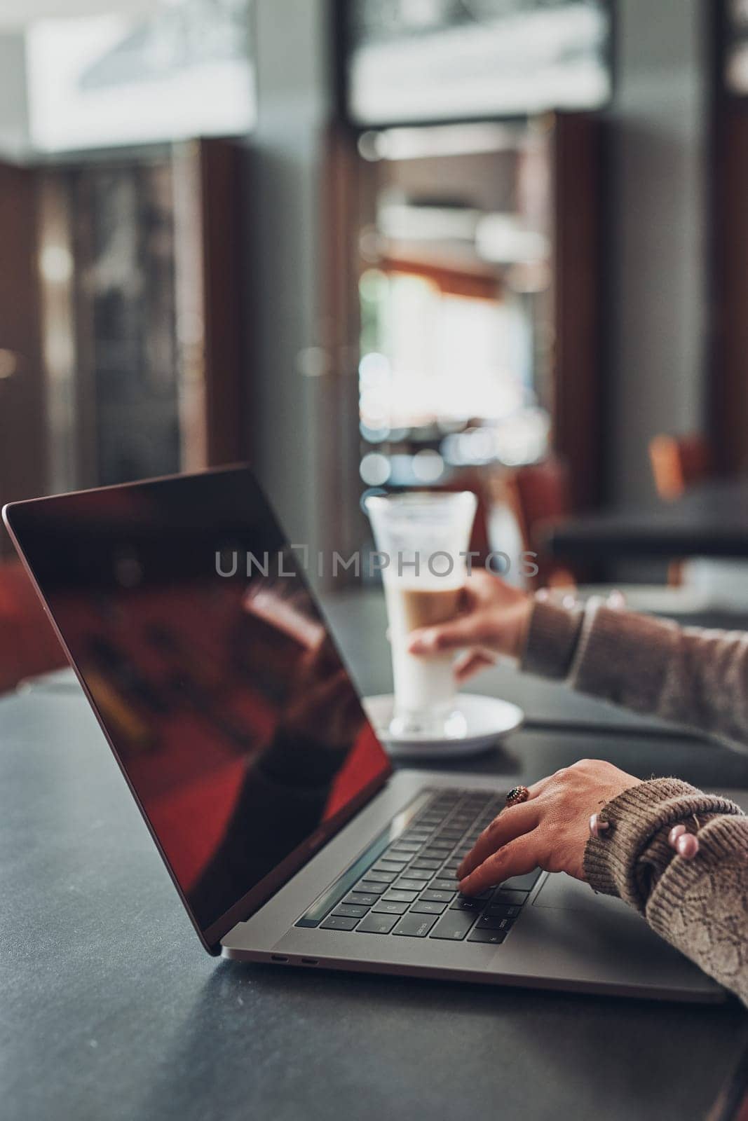 Woman working remotely on her laptop computer managing her work sitting at the table in a cafe. Female hands typing on laptop keyboard