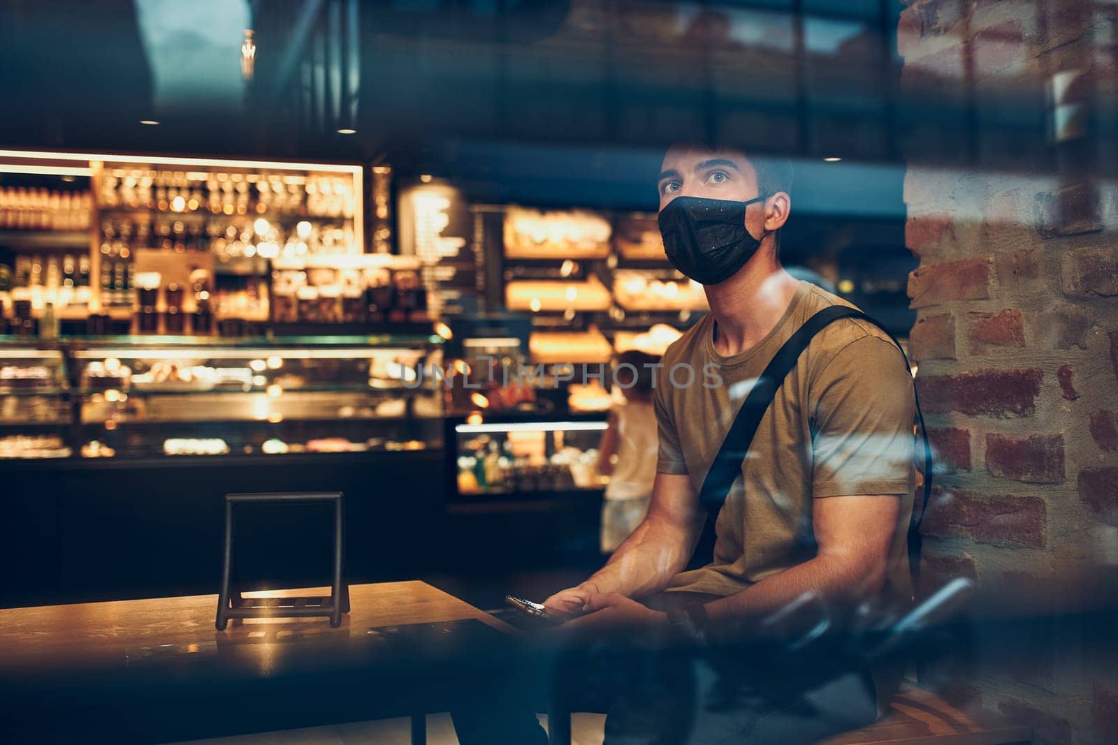Young man sitting in coffee shop at store front in the city center in the evening, wearing the face mask to avoid virus infection and to prevent the spread of disease in time of coronavirus
