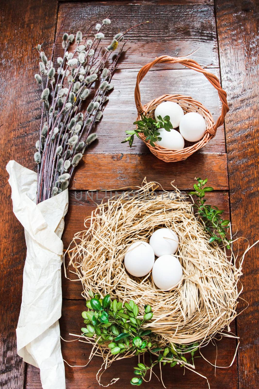 Easter composition of willow twigs with catkins, boxwood, eggs in a easter basket and straw nest, set on old wooden table. Catkins are wrapped up in paper. Inside top shot. Vertical image.
