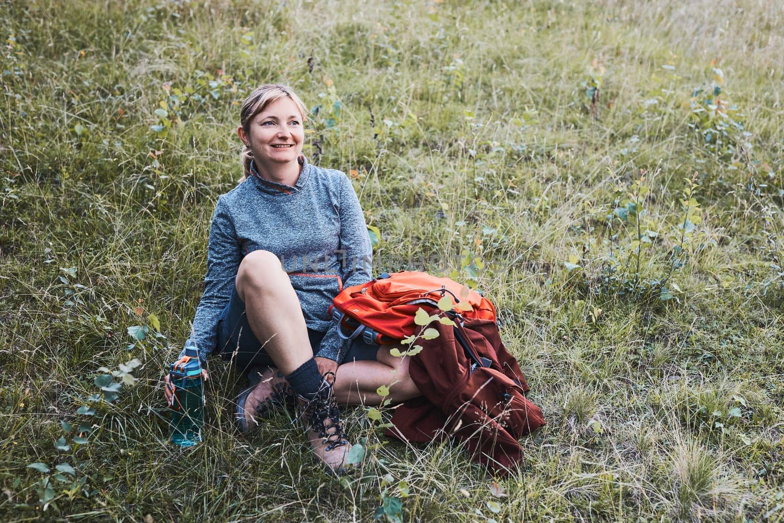 Smiling happy woman with backpack resting on grass during a hike in the mountains, actively spending summer vacation