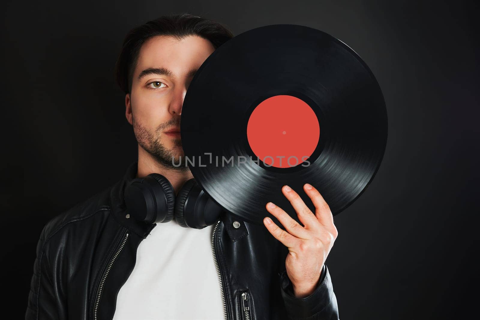 Man holding retro vinyl record covering face. Rock style. Vintage music style. Male wearing black jacket holding vinyl record disc standing on dark background. Retro music. Classic audio