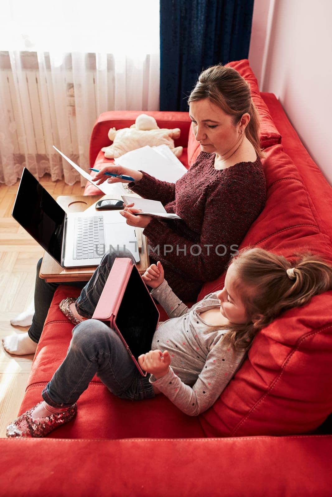 Woman mother working doing her job remotely during video chat call stream online course webinar on laptop from home during COVID-19 quarantine while her daughter watching video on tablet. Woman sitting on bed in front of computer looking at screen