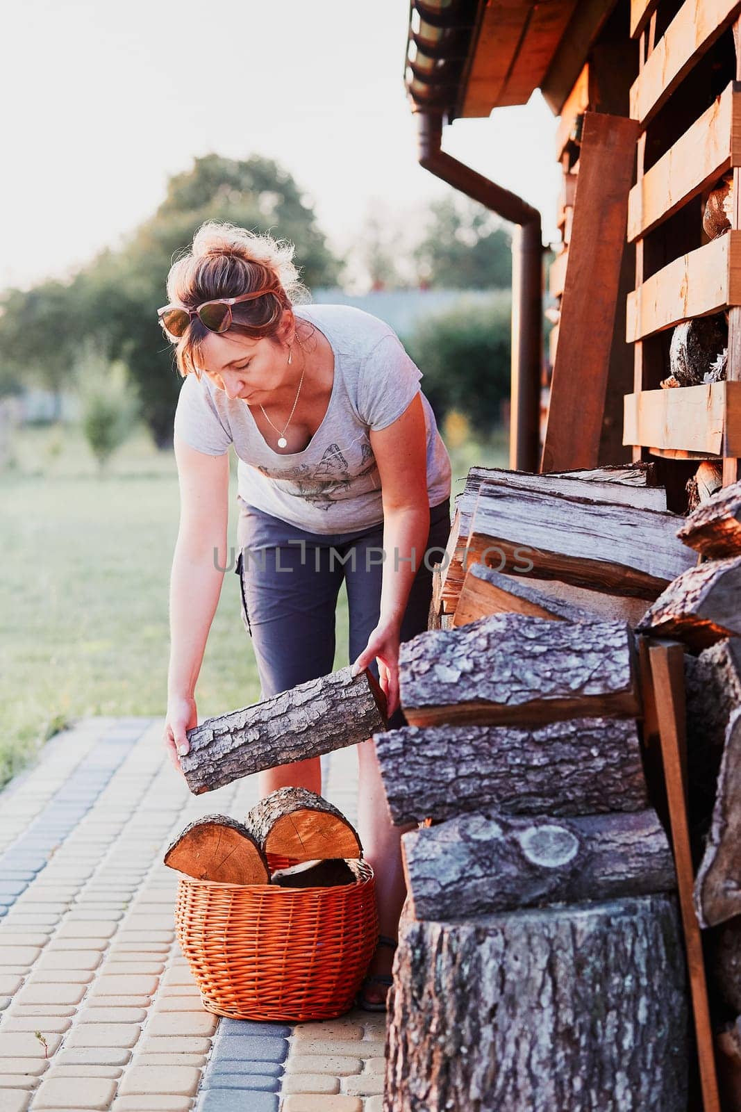 Adult woman putting the wood logs to wicker basket, standing by wood shed in the backyard. Real people, authentic situations