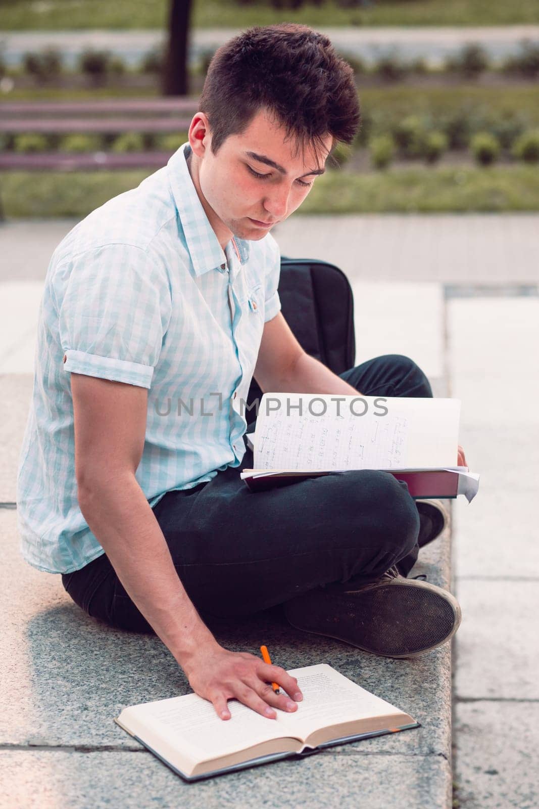Student making the notes learning from books sitting on a monument outside of university. Young boy wearing a blue shirt and dark jeans