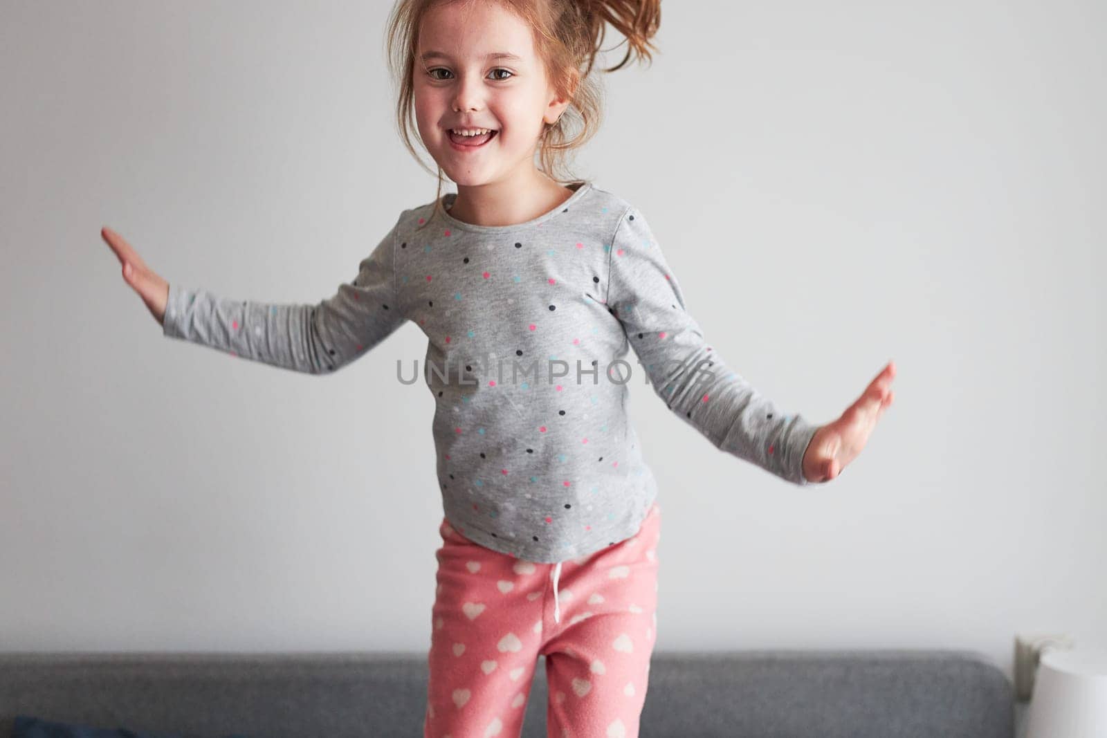 Little happy playful kid girl having fun making silly funny faces jumping on bed in bedroom in the morning at home