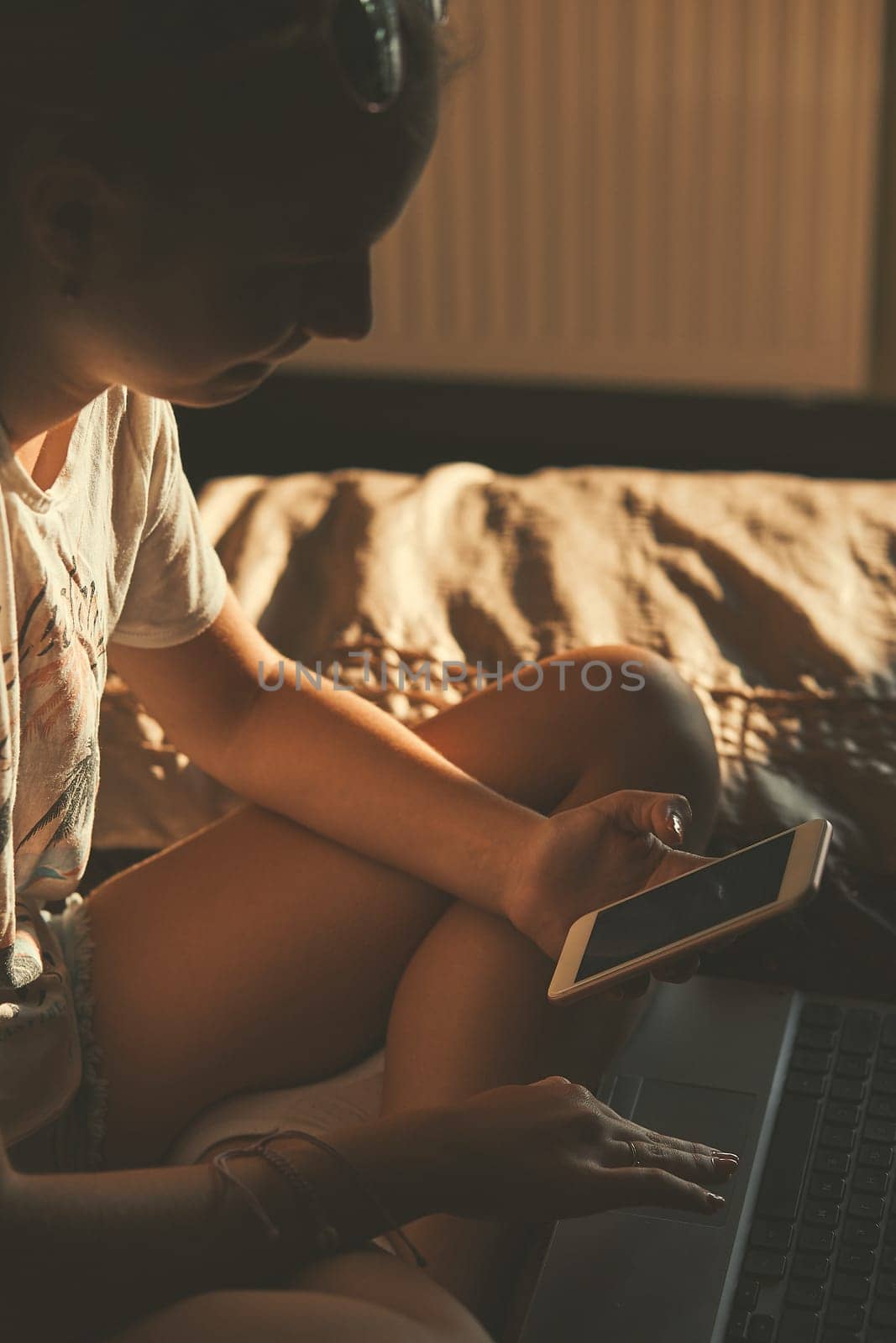 Young woman using portable computer and mobile phone, sitting on a bed, learning online at home. Candid people, real moments, authentic situations