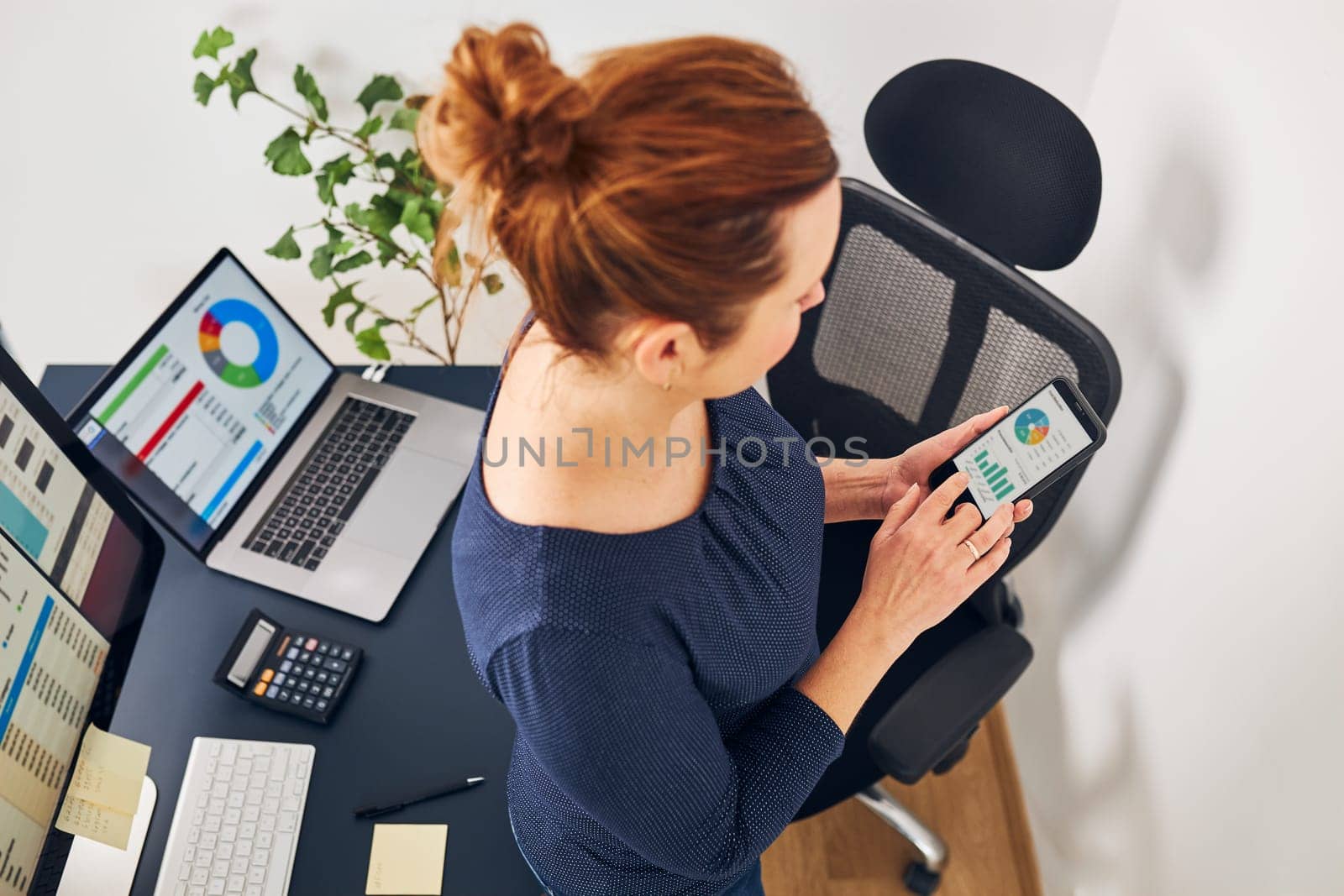 Businesswoman working with financial data on charts and tables on smartphone and computer. Woman entrepreneur standing by desk in office