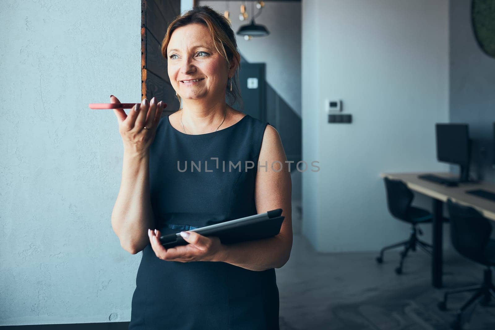 Businesswoman having business call working on tablet in office. Mature busy woman using touch pad computer standing by window in modern interior. Female manager focused on work holding digital device. Using technology