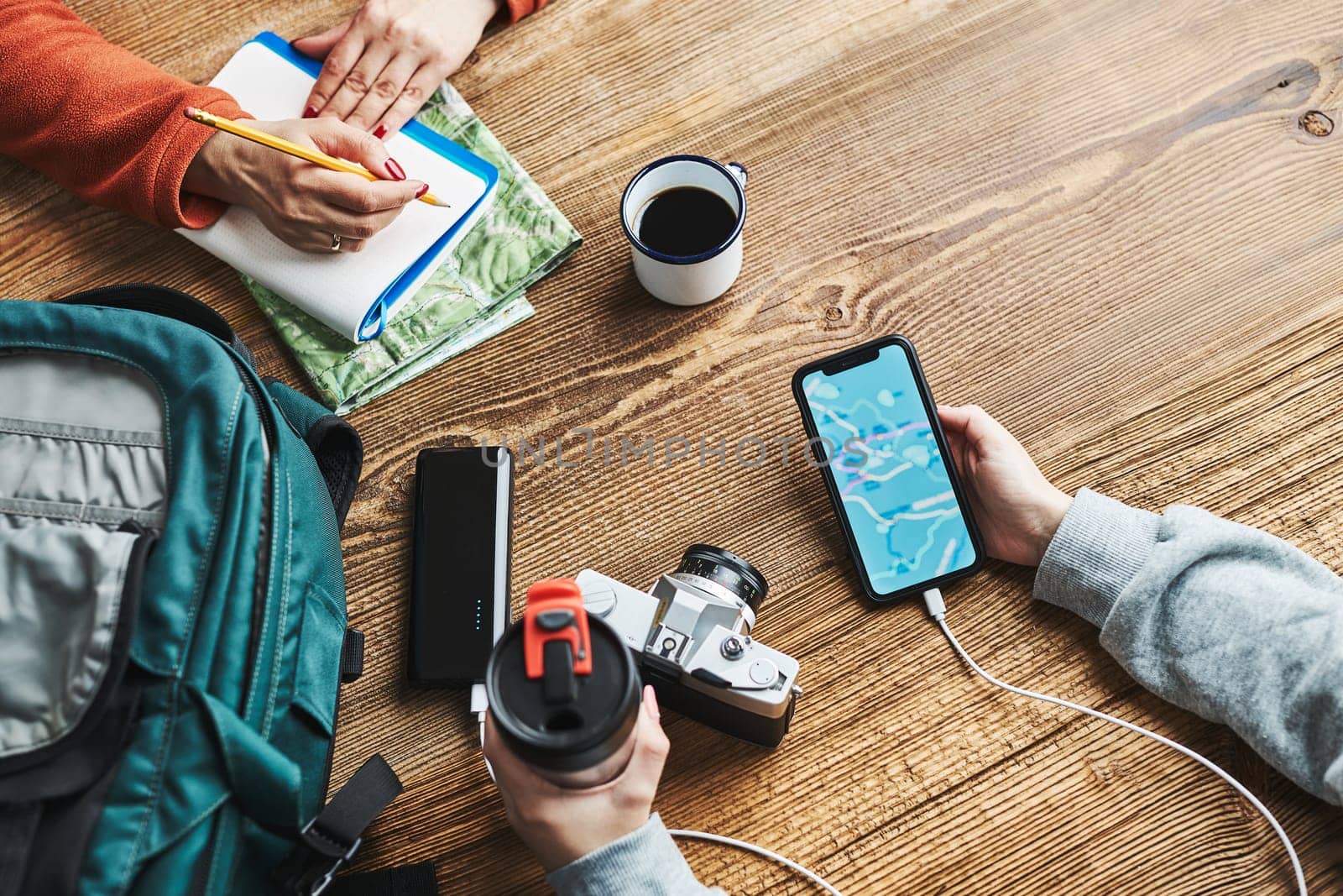 Family planning vacation trip. Girl searching travel destination and routes using navigation map on mobile phone. Woman writing journal and making notes. Charging smartphone with power bank. Using technology while travelling. People sitting at wooden table with camera and backpack during journey. Travel plan concept