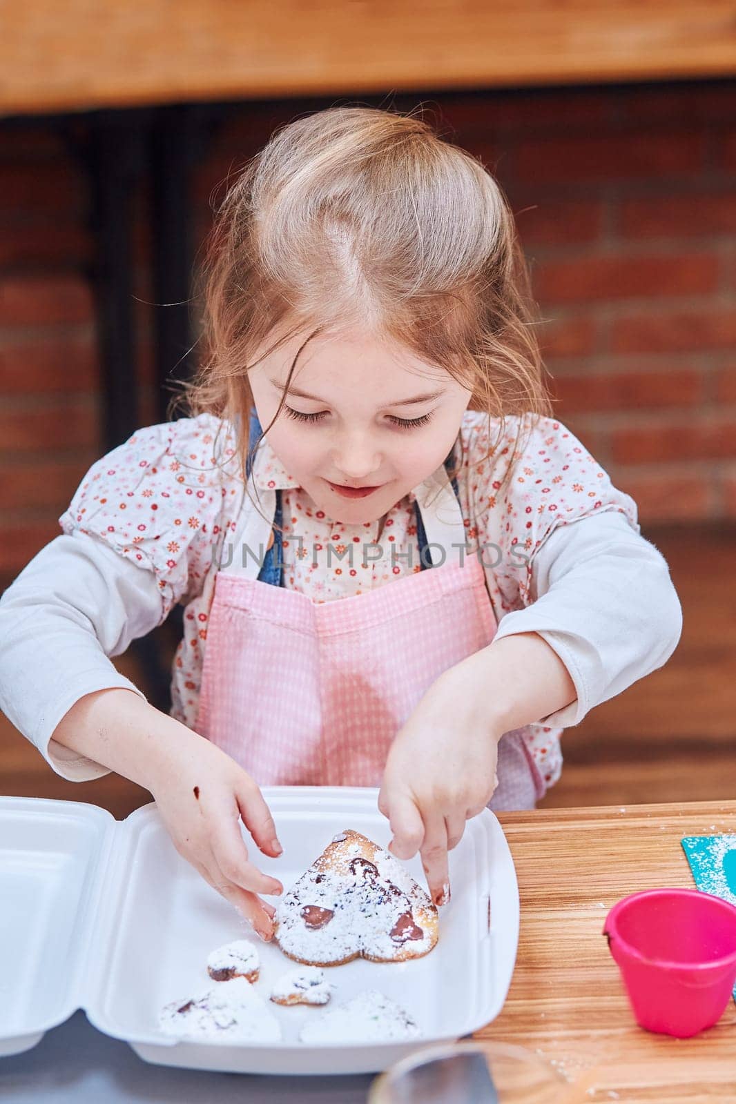 Little girl packing her baked cookies to a box. Kid taking part in baking workshop. Baking classes for children, aspiring little chefs. Learning to cook. Combining and stirring prepared ingredients. Real people, authentic situations